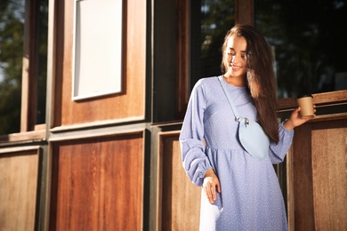 Photo of Beautiful young woman in stylish blue dress with handbag and cup of coffee near wooden building outdoors