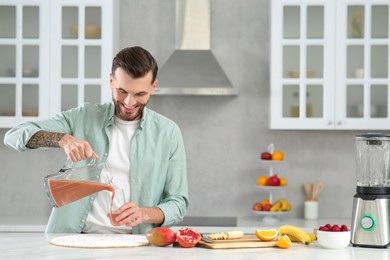 Handsome man pouring tasty smoothie into glass at white marble table in kitchen. Space for text