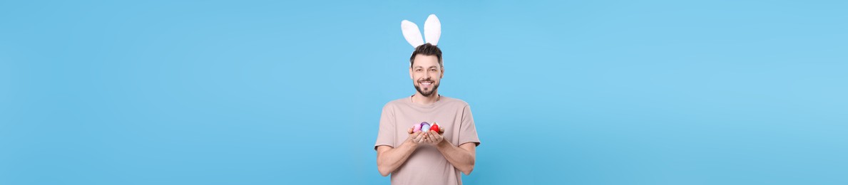 Happy man with bunny ears holding Easter eggs on light blue background. Banner design