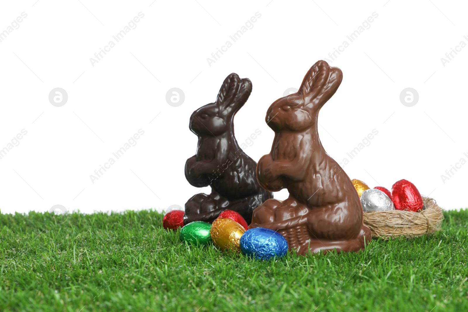 Photo of Chocolate bunnies and eggs on green grass against white background, space for text. Easter celebration