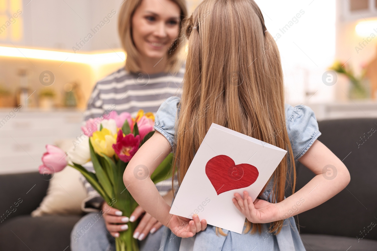 Photo of Little girl hiding greeting card for mom at home, selective focus. Happy Mother's Day