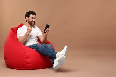 Photo of Happy man with smartphone sitting on bean bag against light brown background. Space for text