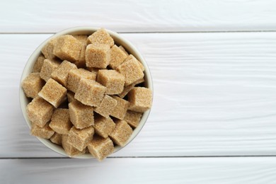 Photo of Brown sugar cubes in bowl on white wooden table, top view. Space for text