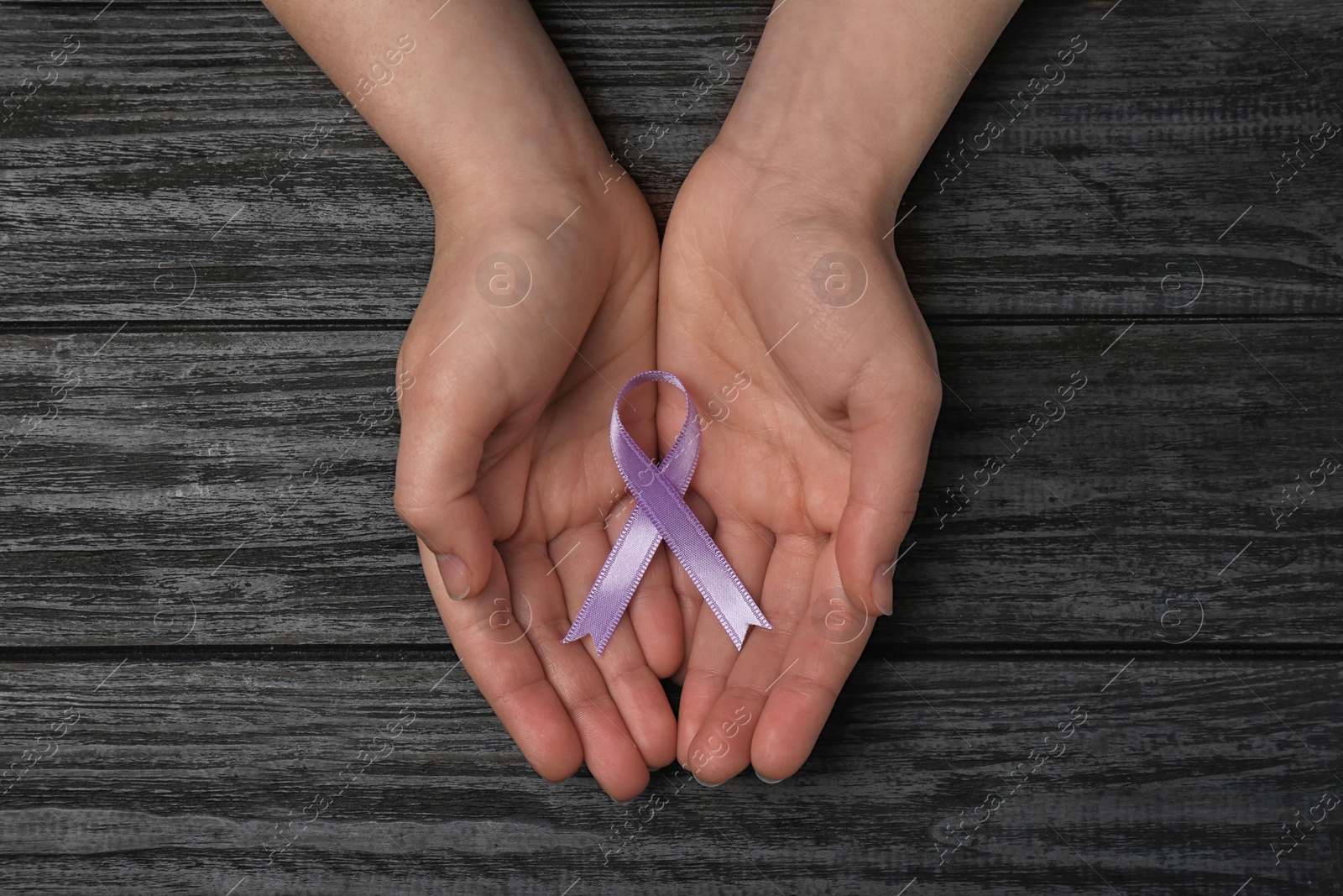 Photo of Woman holding purple awareness ribbon on black wooden background, top view