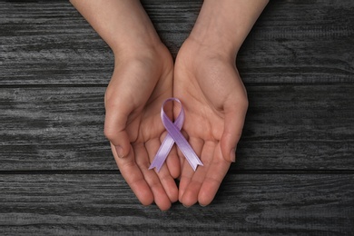 Woman holding purple awareness ribbon on black wooden background, top view