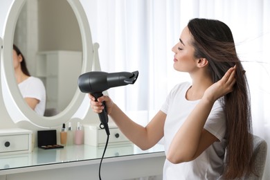 Photo of Beautiful young woman using hair dryer near mirror at home