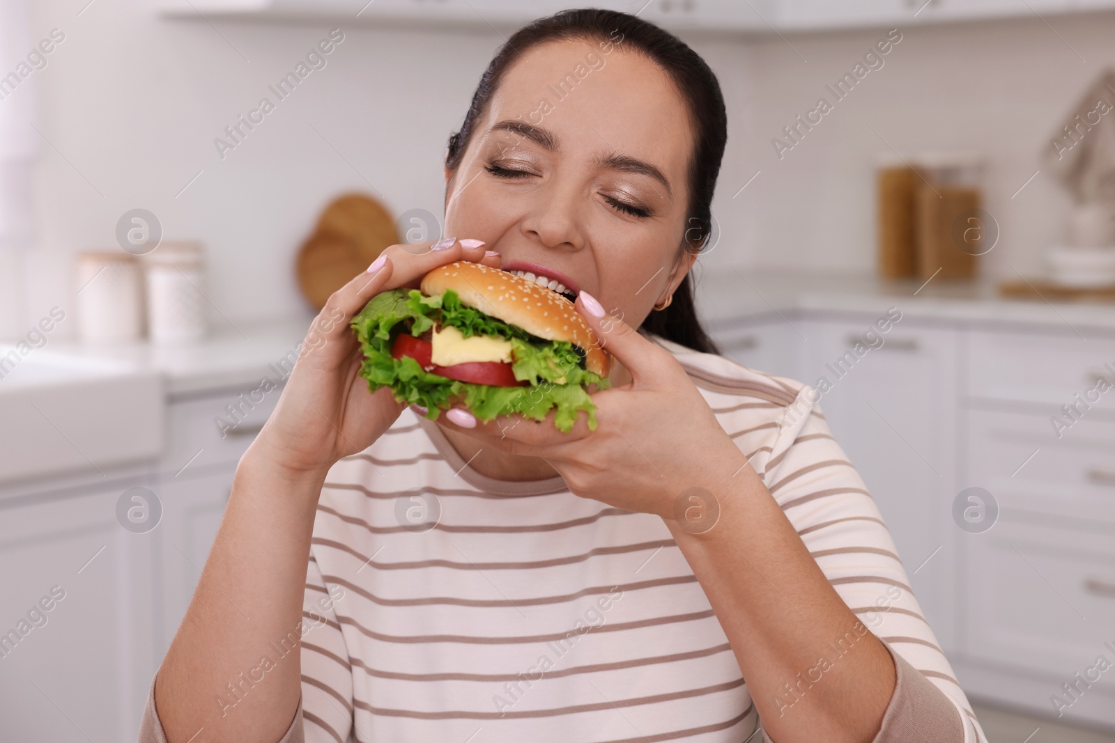 Photo of Overweight woman eating burger in kitchen at home