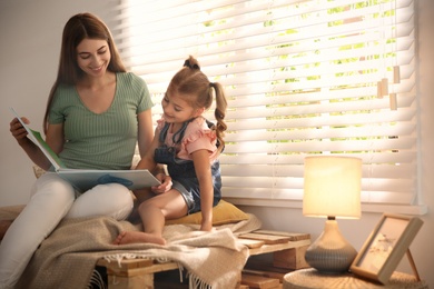 Photo of Young woman and her daughter reading book near window at home