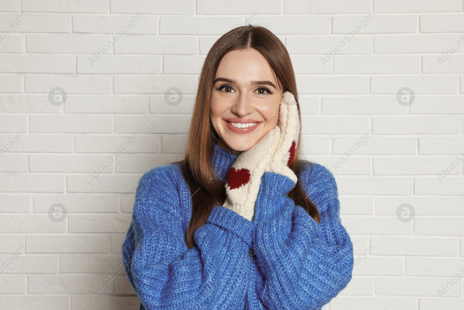 Photo of Beautiful young woman in mittens and blue sweater near white brick wall. Winter season