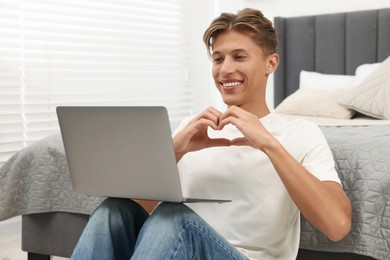 Happy young man having video chat via laptop and making heart with hands indoors. Long-distance relationship