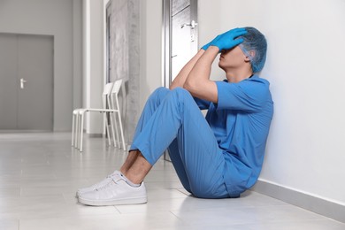 Photo of Exhausted doctor sitting near grey wall in hospital hallway