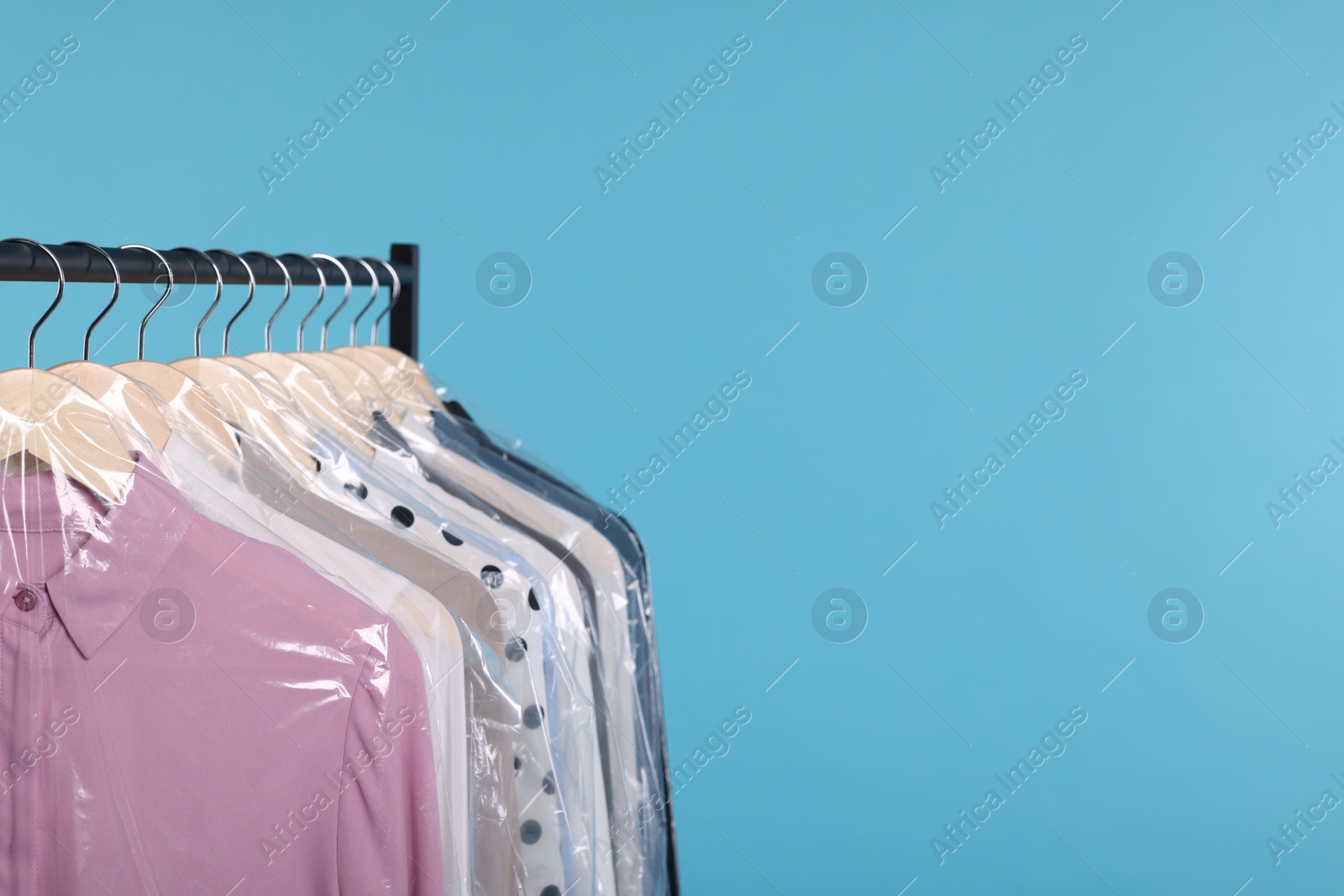 Photo of Dry-cleaning service. Many different clothes in plastic bags hanging on rack against light blue background, space for text