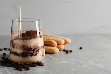 Tasty tiramisu in glass, coffee beans, spoon and biscuits on light grey table, closeup. Space for text