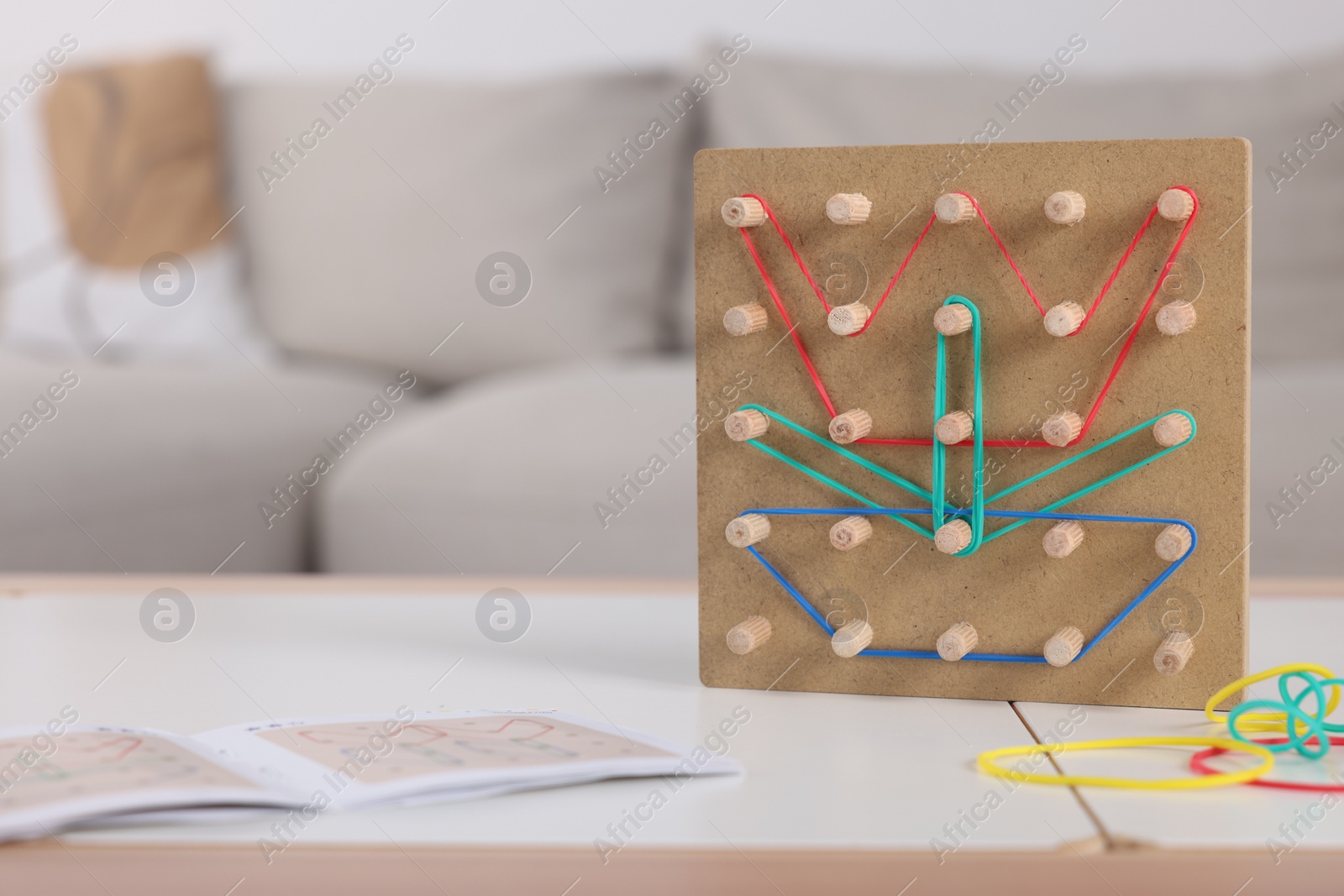 Photo of Wooden geoboard with flower made of rubber bands and activity book on white table in room, space for text. Motor skills development