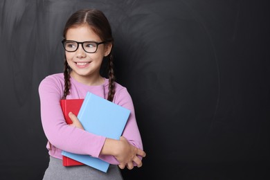 Cute schoolgirl in glasses holding books near chalkboard, space for text