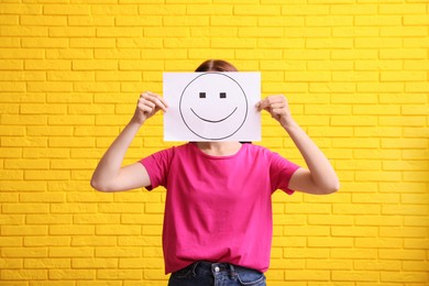 Woman hiding behind sheet of paper with happy face against yellow brick wall