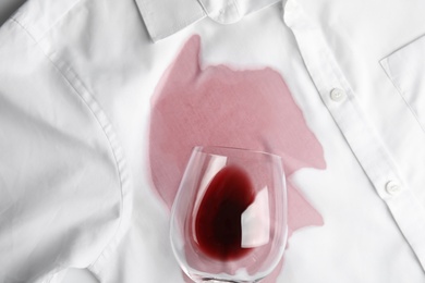 Photo of Overturned glass and spilled exquisite red wine on white shirt, top view