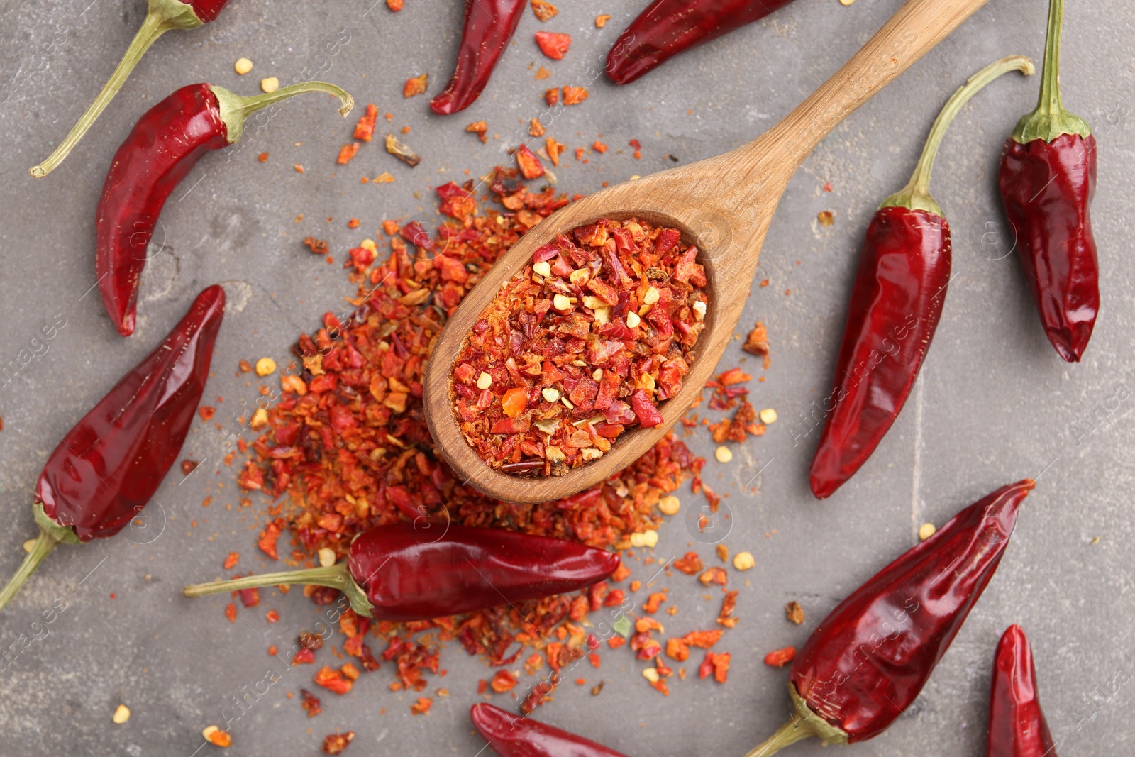 Photo of Chili pepper flakes, pods and spoon on grey table, flat lay