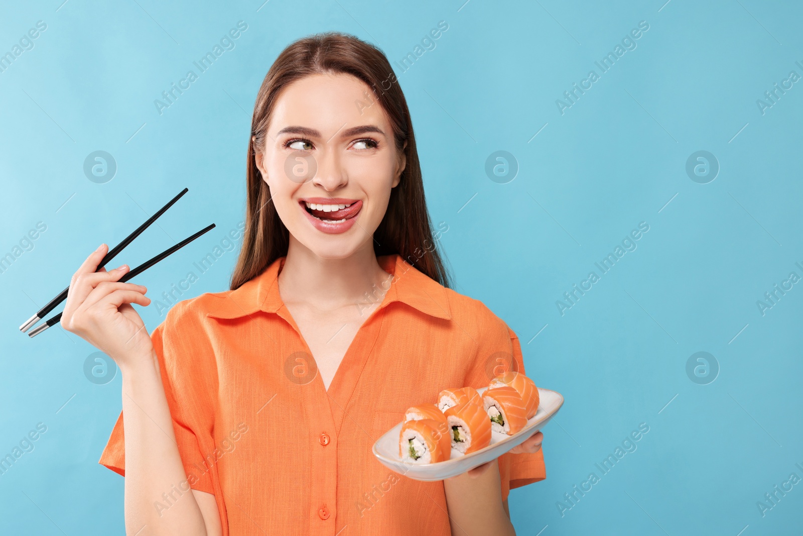 Photo of Happy young woman with plate of sushi rolls and chopsticks on light blue background. Space for text