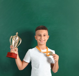 Happy boy with golden winning cup and medal near chalkboard