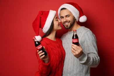 Photo of MYKOLAIV, UKRAINE - JANUARY 27, 2021: Young couple in Christmas hats holding bottles of Coca-Cola on red background
