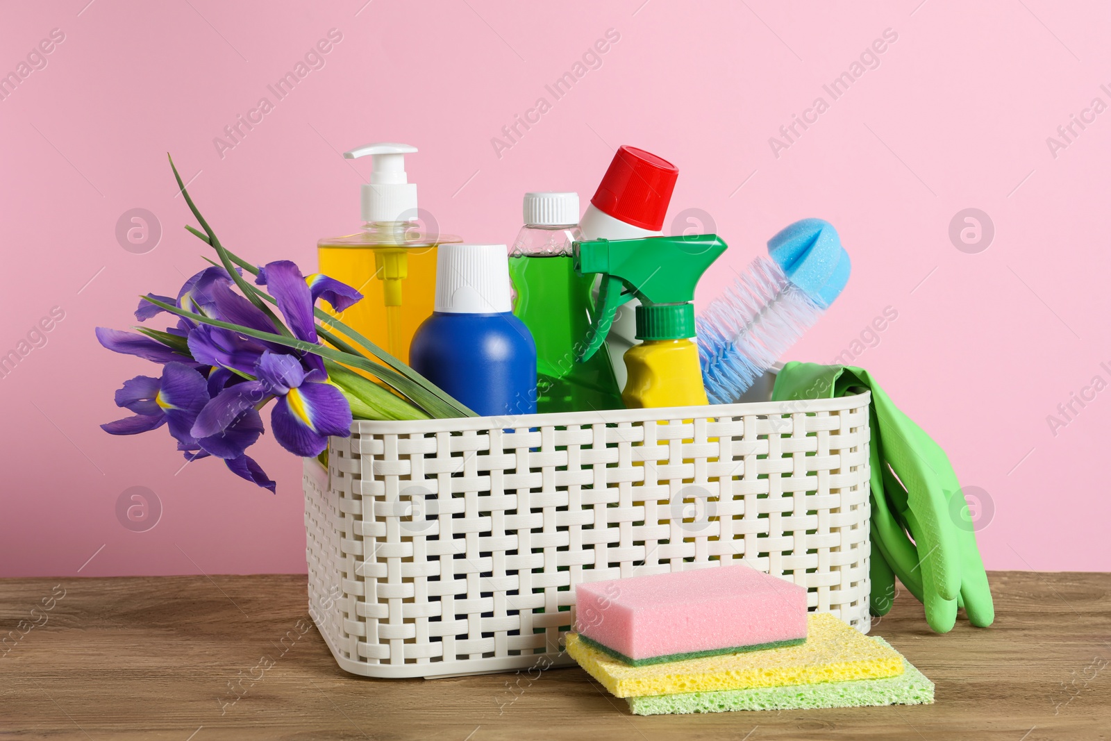 Photo of Spring cleaning. Basket with detergents, flowers and sponges on wooden table against pink background