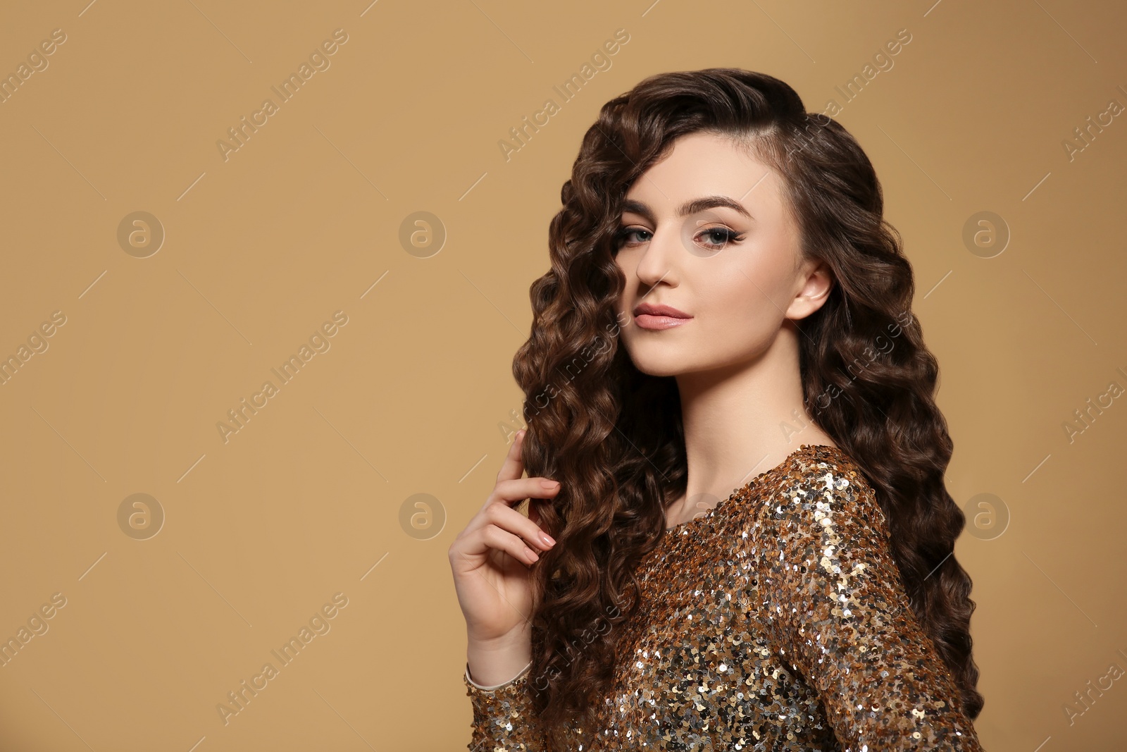 Photo of Beautiful young woman with long curly brown hair in golden sequin dress on beige background, space for text