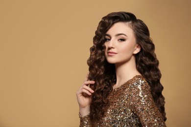 Beautiful young woman with long curly brown hair in golden sequin dress on beige background, space for text