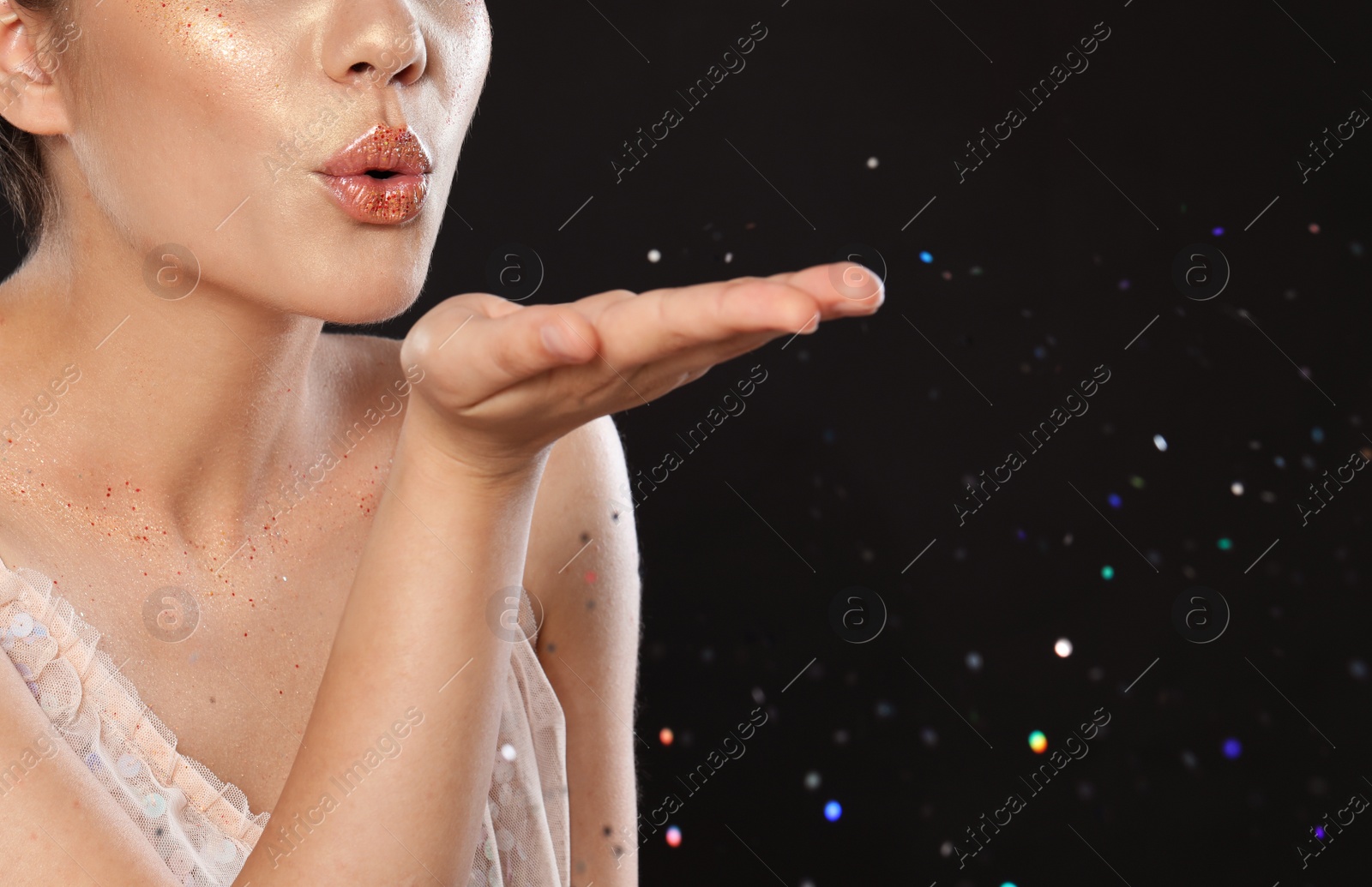Photo of Beautiful young woman with glitter makeup blowing kiss on black background, closeup view