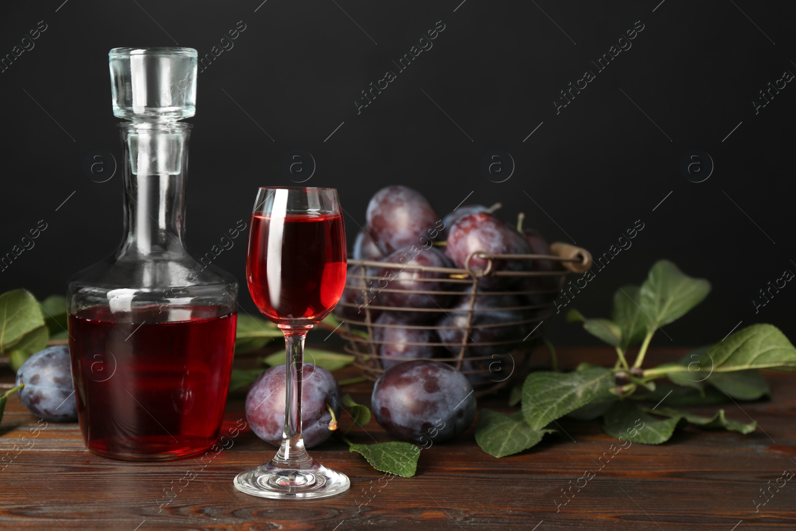 Photo of Delicious plum liquor and ripe fruits on wooden table against black background. Homemade strong alcoholic beverage