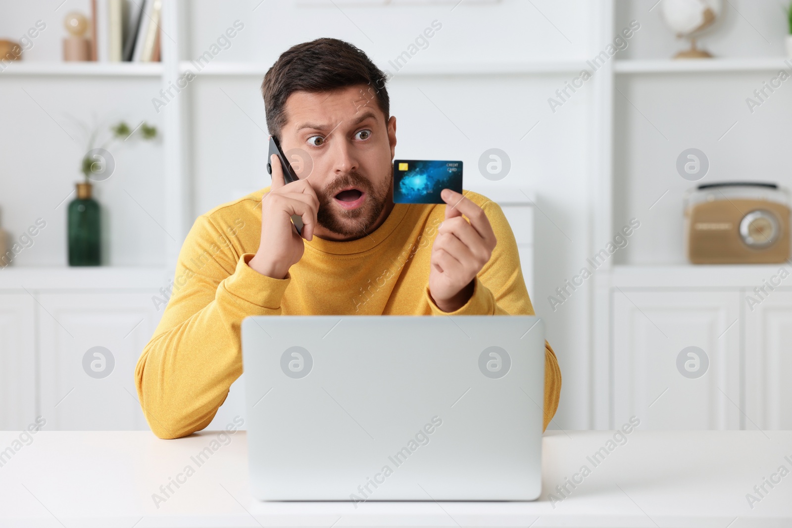 Photo of Scared man with credit card talking on smartphone near laptop at home. Be careful - fraud
