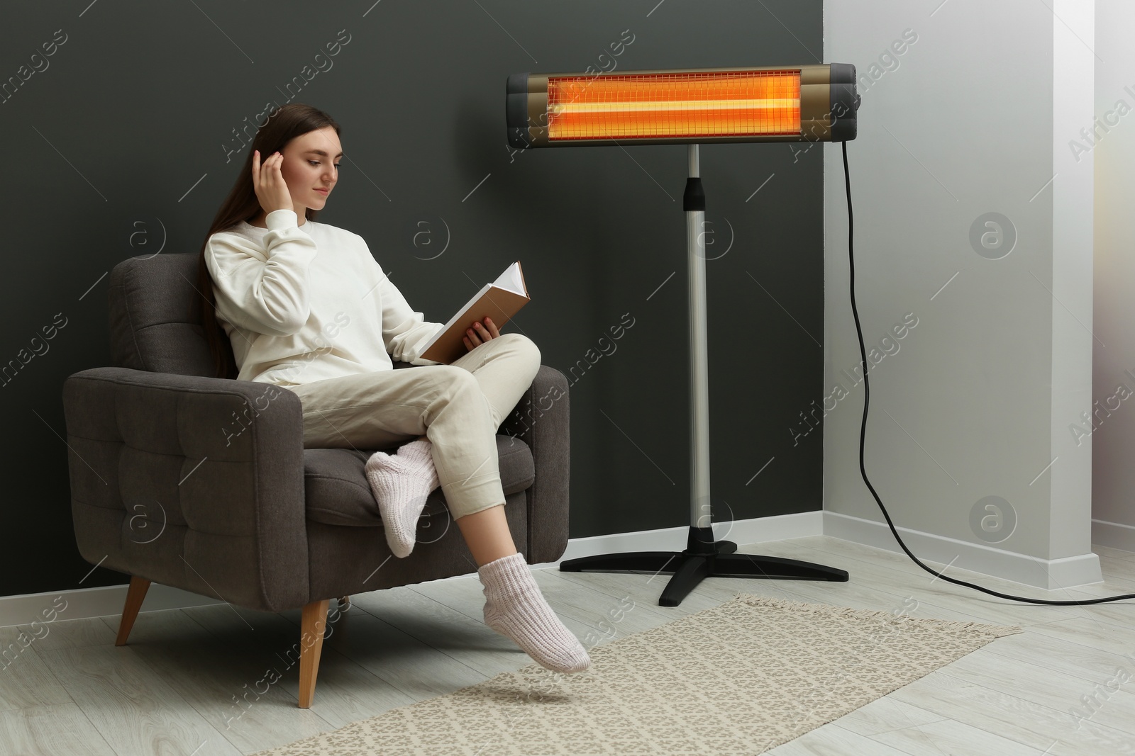 Photo of Young woman reading book on armchair near electric infrared heater at home. Space for text