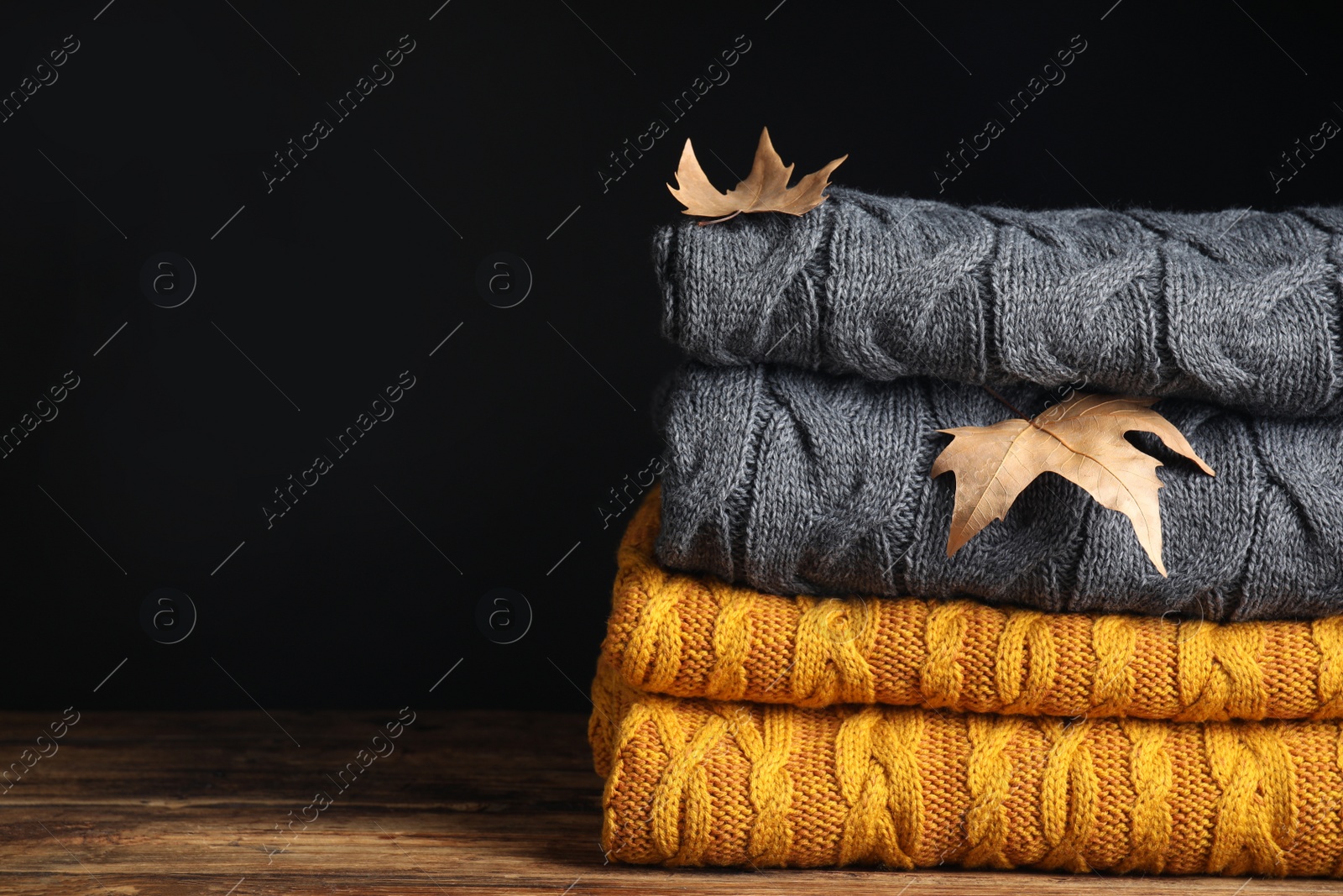 Photo of Grey and orange knitted plaids with dry leaves on wooden table, closeup. Space for text