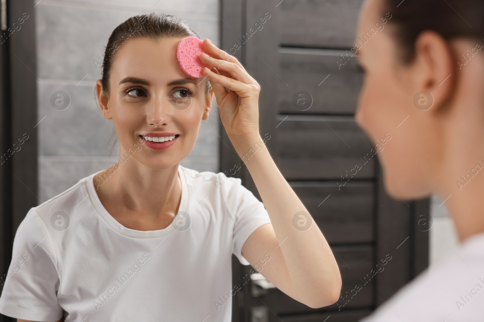 Photo of Happy young woman washing her face with sponge near mirror in bathroom