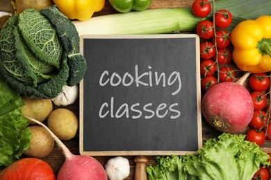 Small chalkboard with inscription Cooking Classes and different fresh vegetables on wooden table, flat lay