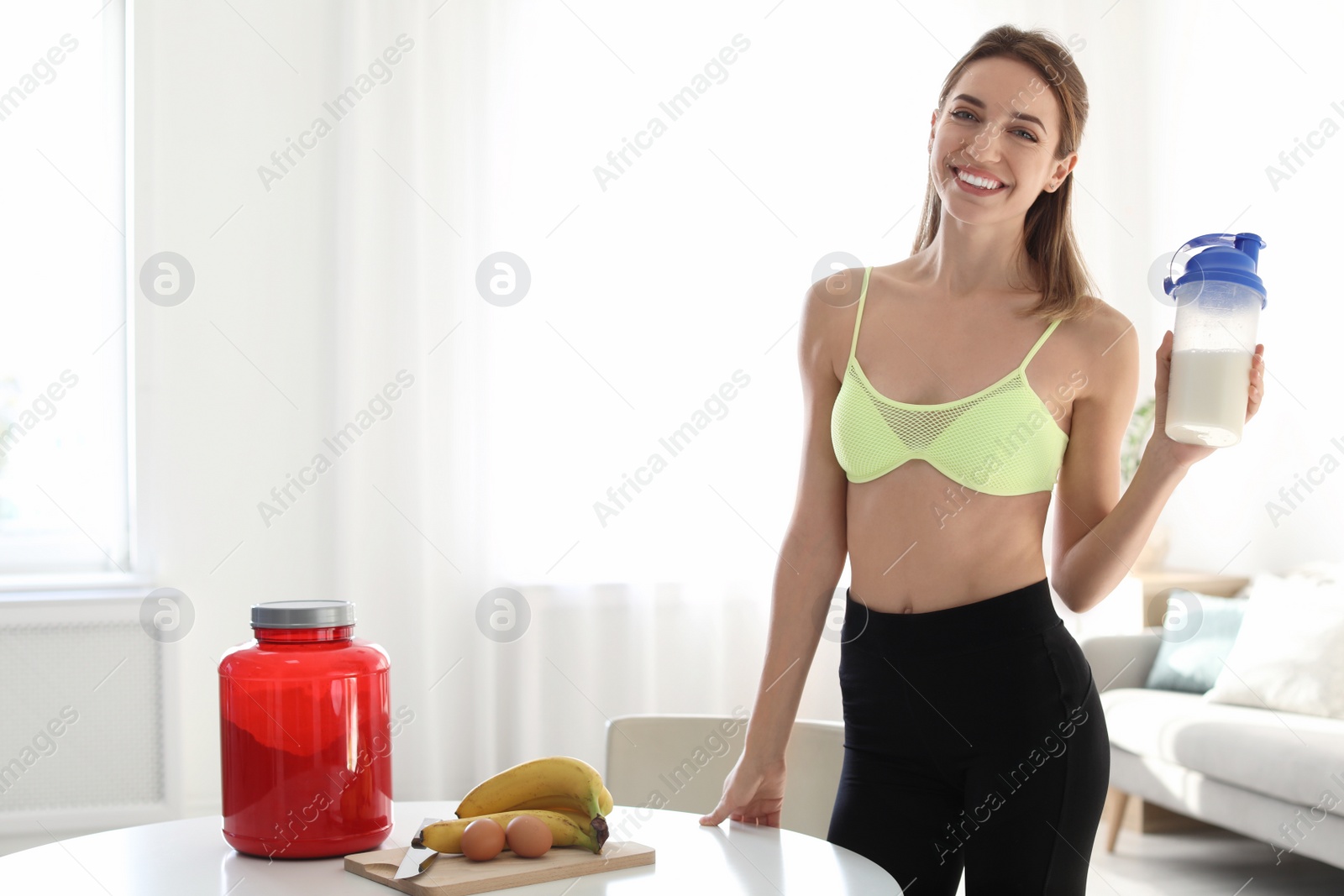 Photo of Young woman holding bottle of protein shake near table with ingredients in room. Space for text