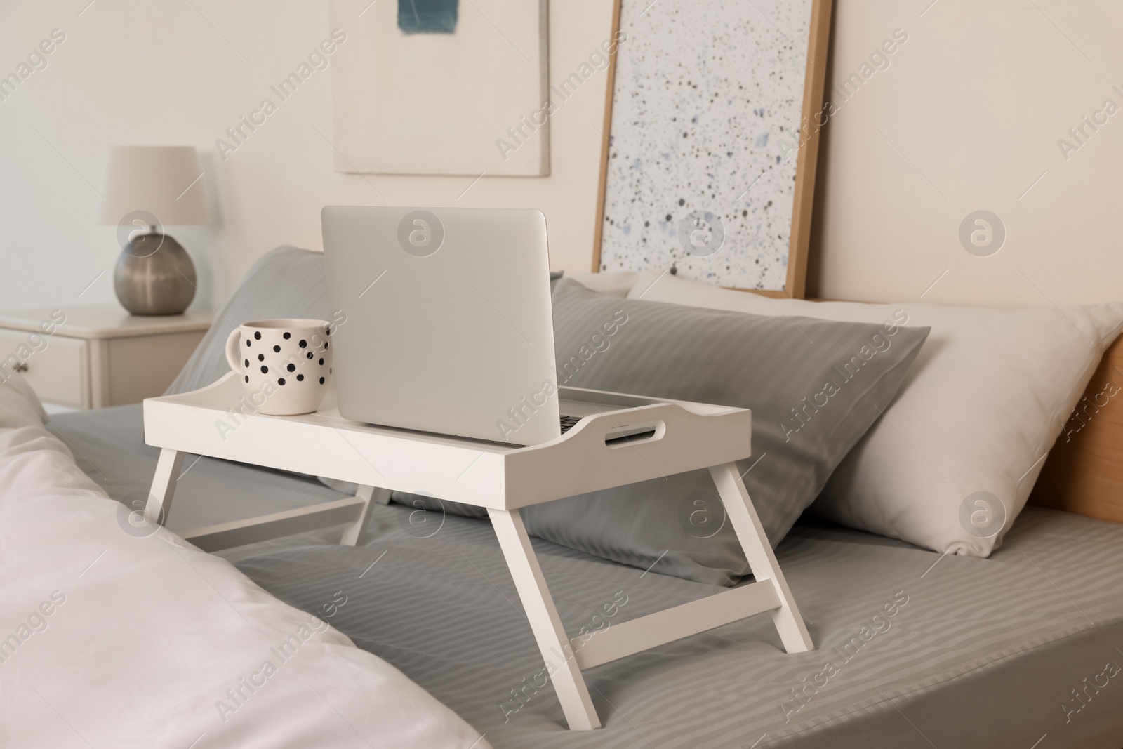 Photo of White tray table with laptop and cup of drink on bed indoors