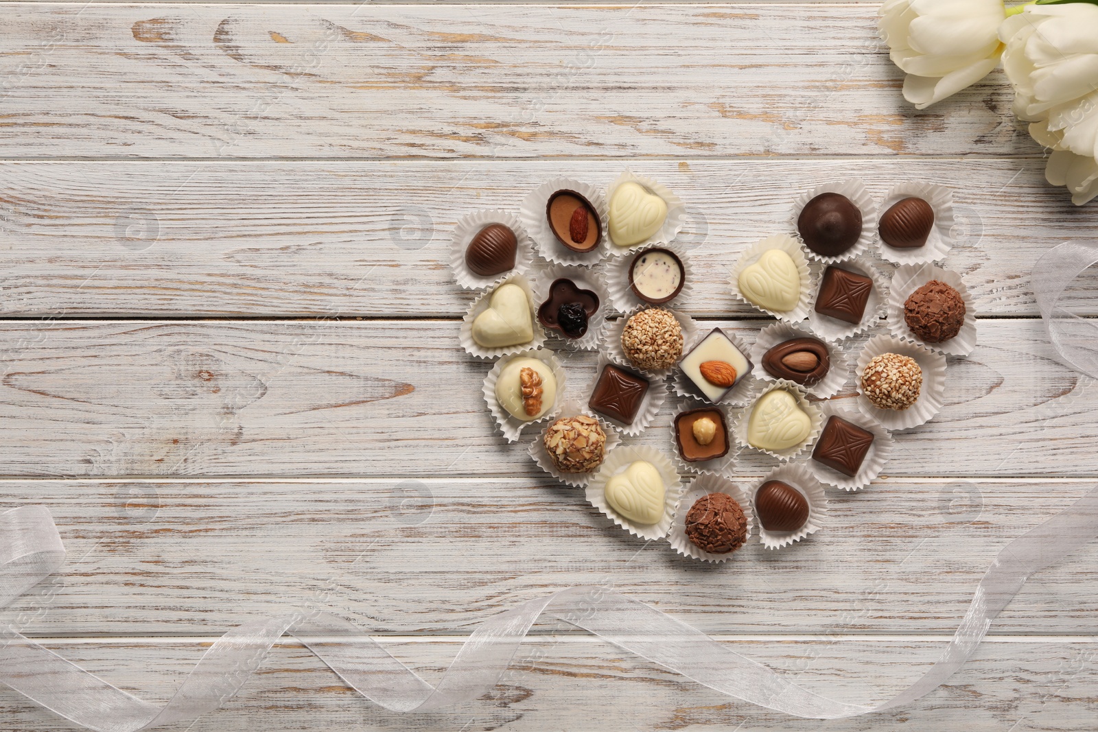 Photo of Heart made with delicious chocolate candies and beautiful tulips on white wooden table, flat lay. Space for text