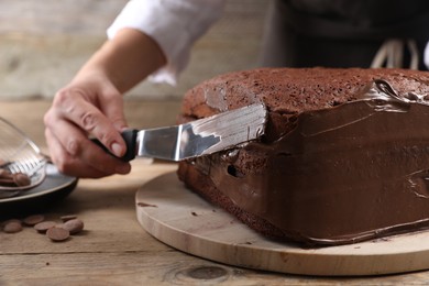 Photo of Woman smearing sides of sponge cake with chocolate cream at wooden table, closeup