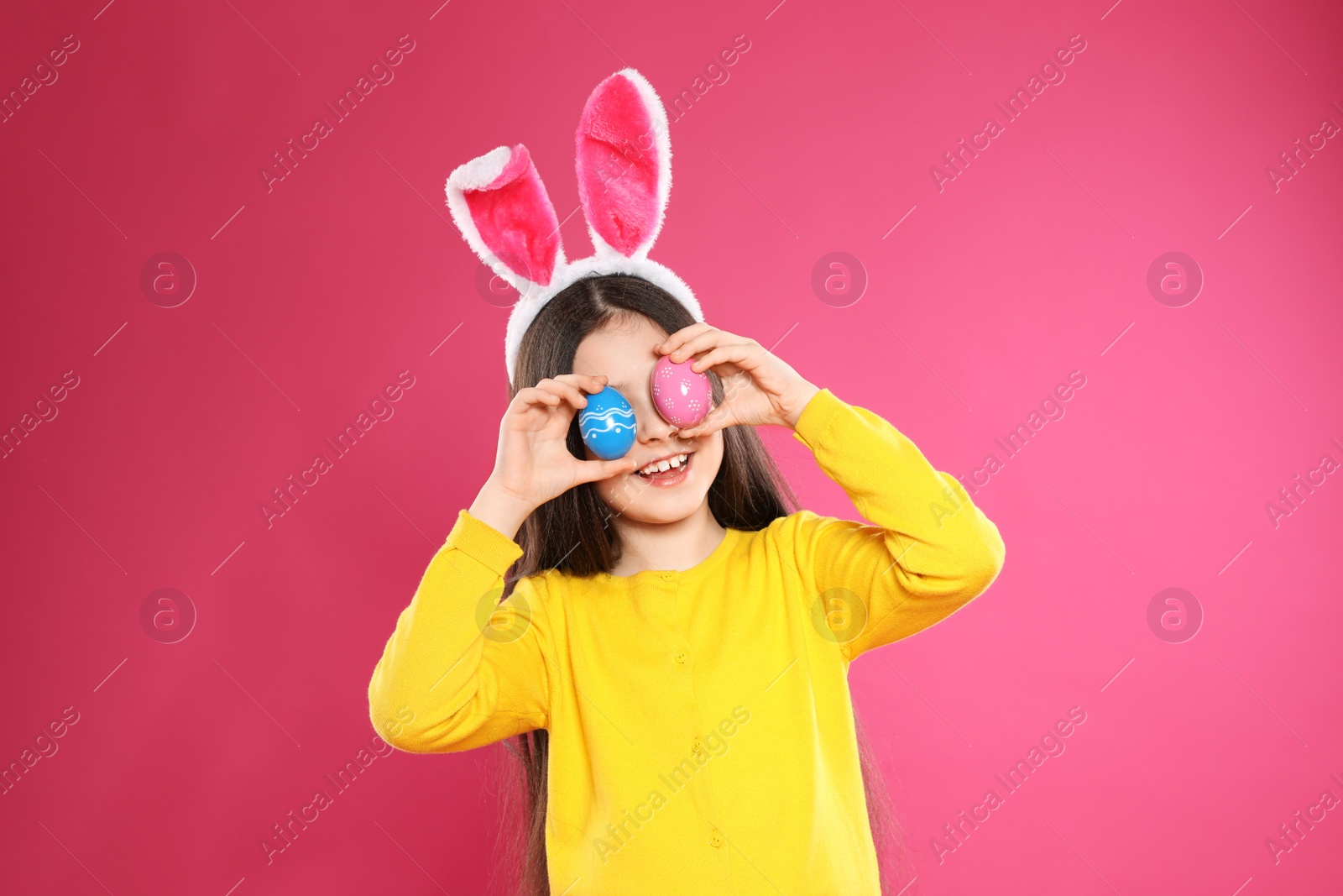 Photo of Little girl in bunny ears headband holding Easter eggs near eyes on color background