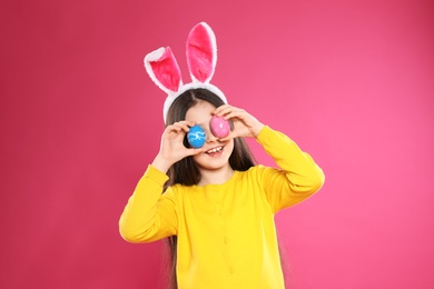 Little girl in bunny ears headband holding Easter eggs near eyes on color background