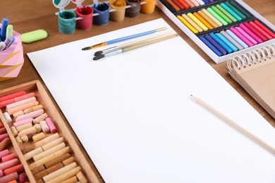 Photo of Blank sheet of paper, colorful chalk pastels and other drawing tools on wooden table. Modern artist's workplace