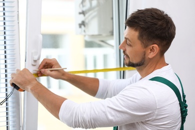 Handyman with tape measure and pencil installing window blinds indoors