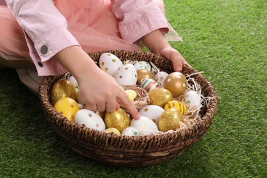 Photo of Child with wicker basket full of Easter eggs on green grass, closeup