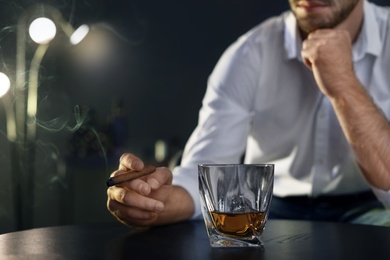 Man with glass of whiskey and cigar sitting at table, closeup. Space for text