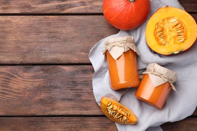 Photo of Jars of pumpkin jam and fresh pumpkin on wooden table, flat lay. Space for text