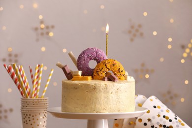 Photo of Delicious cake decorated with sweets and burning candle against blurred lights
