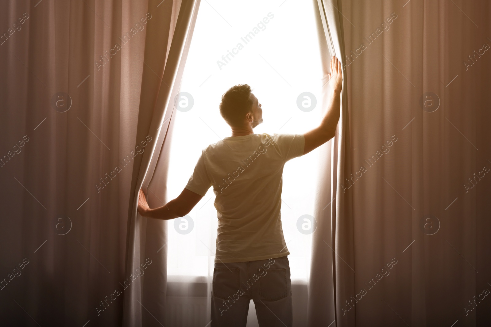 Photo of Man opening window curtains at home, back view