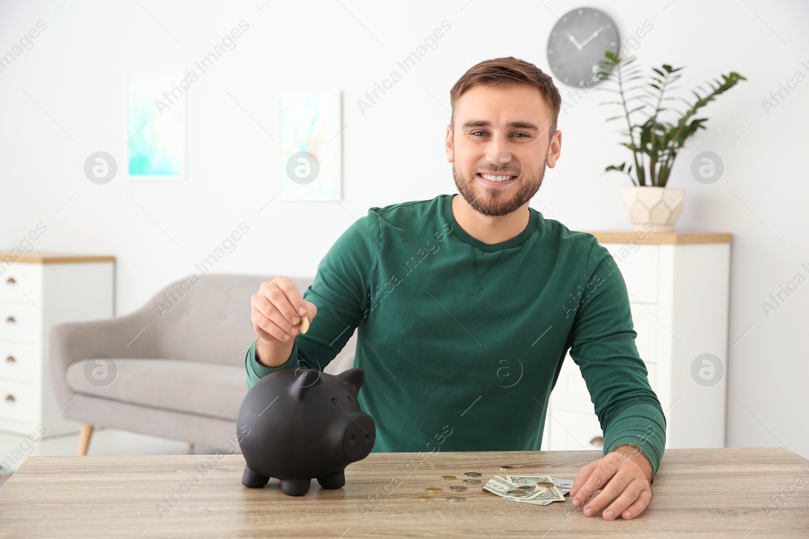 Photo of Young man putting coin into piggy bank at table indoors