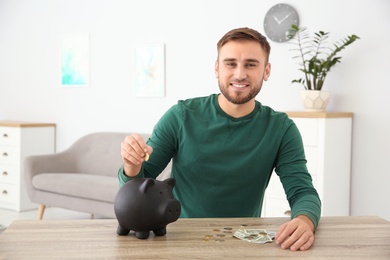 Young man putting coin into piggy bank at table indoors